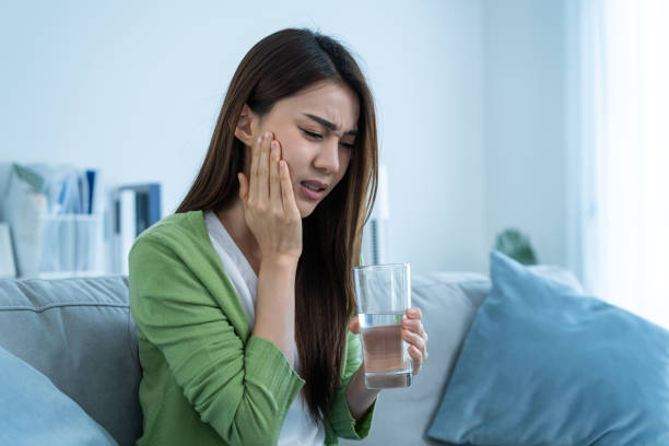 mulher asiática bonita sente dor de dente terrível depois de beber água fria. mulheres atraentes sentam-se no sofá na sala tocando a bochecha, sentem-se magoados e sofrendo de dor de dente sensível, dor e cáries. - dor de dente - fotografias e filmes do acervo