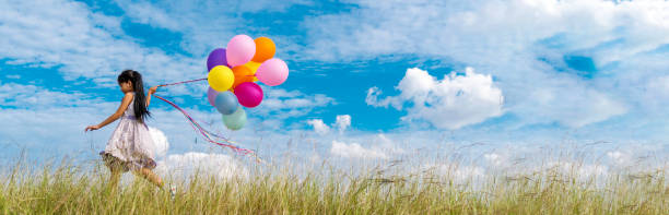 banner cheerful cute girl holding balloons running green meadow white cloud and blue sky with happiness. panorama hands holding vibrant air balloons happy times summer sunlight outdoor copy space - grass area field air sky imagens e fotografias de stock