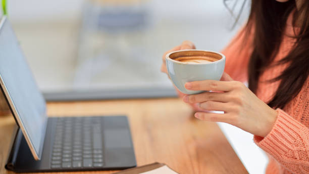 istock Young female in pink sweater drinking, holding hot coffee cup while working on tablet 1347406887