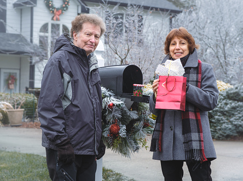 Smiling senior couple looking at camera while standing by their mailbox on snowy winter day; senior woman holding red bag with Christmas presents
