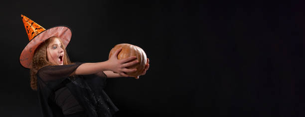 Playful girl in witch costume holds pumpkin with frightened expression stock photo