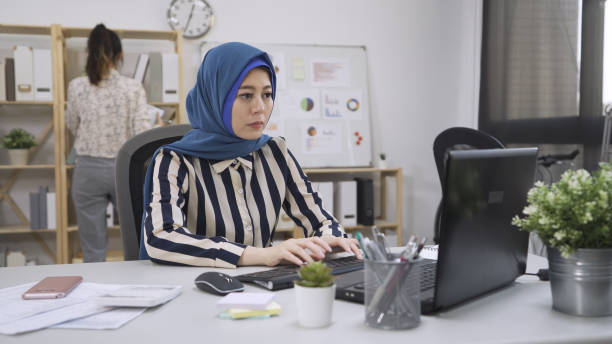 muslim woman connected on internet with laptop typing on keyboard replying emails to client order. japanese colleague standing in background gind document files in bright cozy office. multi ethnic. mujer musulmana conectada a internet con computadora portátil escribiendo en el teclado respondiendo correos electrónicos a la orden del cliente. colega japonés de pie en los archivos de documentos gind de fondo en una oficina luminosa y acogedora. mult