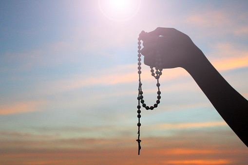 Silhouette of a hand holding Rosary on colorful sunset sky light background. Person with Rosary in hands with Jesus Christ crucifix. Copy space for spiritual text message design. Catholic symbol.