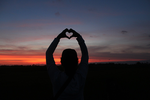 Silhouette of a person holding a heart shaped hands. Person making love sign against colorful romantic sunset sunrise sky clouds backgrounds. Love background.