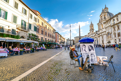 Rome, Italy, October 13 -- A street artist awaits some customers in Piazza Navona, in the historic and baroque heart of Rome. On the right, the majestic facade of the church of Sant'Agnese, a baroque masterpiece by the architect Francesco Borromini, while on the left some typical Italian restaurants along the square. The Rione Parione, the district surrounding Piazza Navona, is one of the most beautiful and visited districts of Rome for the presence of countless artistic and historical treasures, monuments and ancient Romanesque and Baroque churches, but also for its squares and alleys to explore freely, where it is easy to find typical restaurants, small artisan shops, street artists, and the original Roman soul. In 1980 the historic center of Rome was declared a World Heritage Site by Unesco. Image in High Definition format.
