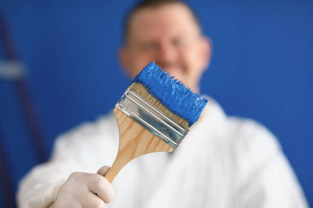A man in uniform holds a paintbrush with blue paint close-up. Painting the wall in the room. Renovation of the premises