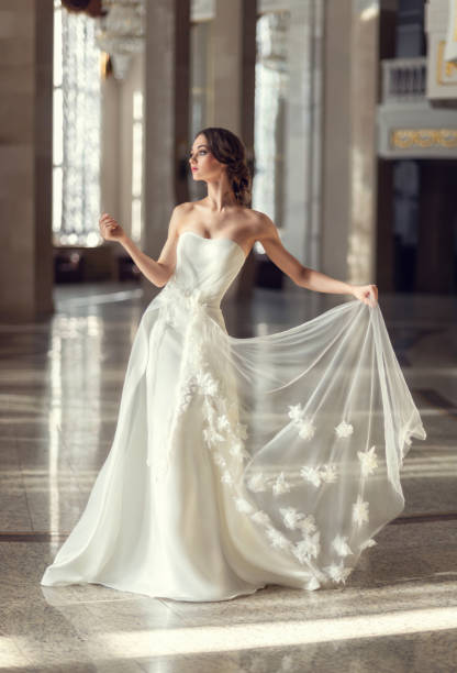 jeune fille brune fragile. mariée dans une robe de mariée luxueuse dans un intérieur chic avec une belle lumière du soleil des fenêtres coiffure de mariage et maquillage. photo pour le sel de mariage, salon de beauté - robe de mariée photos et images de collection