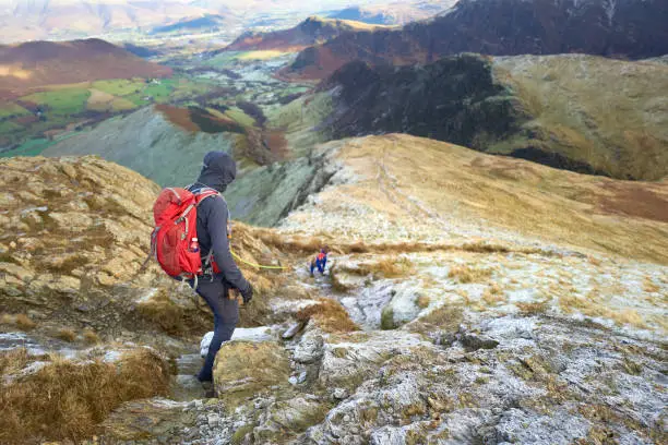 A hiker and their dog descending the frozen frost covered rocky mountain ridge from the summit of Robinson towards High Snab Bank on a cold winters day in the Lake District, UK.