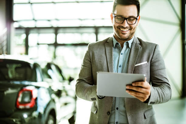 Happy salesman using digital tablet in car showroom. Young happy car salesperson using digital tablet in a showroom. car salesperson photos stock pictures, royalty-free photos & images