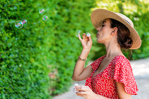 Side view of tranquil female in summer outfit standing in lush garden and blowing soap bubbles while having fun at weekend. Concept have fun like a child
