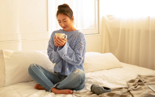 jovem asiática relaxando com xícara de café ou chá curtindo a noite ensolarada em um dia frio de inverno enquanto se senta na cama no quarto. conceito de estilo de vida feminino e inverno. outono, atmosfera de inverno. - warm light - fotografias e filmes do acervo