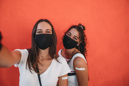 Two young adult women are taking a selfie showing their arms where they have just been vaccinated. They are standing against an orange wall, wearing protective face masks.