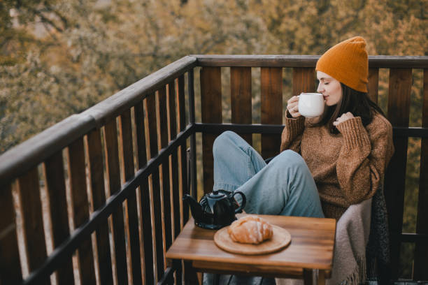 giovane donna in maglia maglione e cappello che beve tè e mangia croissant freschi sull'accogliente balcone di una casa di campagna in legno. - 30s women sweater female foto e immagini stock