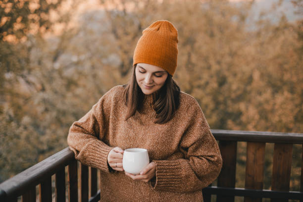 giovane donna in maglia maglione e cappello che beve tè e mangia croissant freschi sull'accogliente balcone di una casa di campagna in legno. - balcony house golf home interior foto e immagini stock