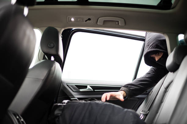 istock Hooded thief with mask stealing backpack from a parked car 1347457686