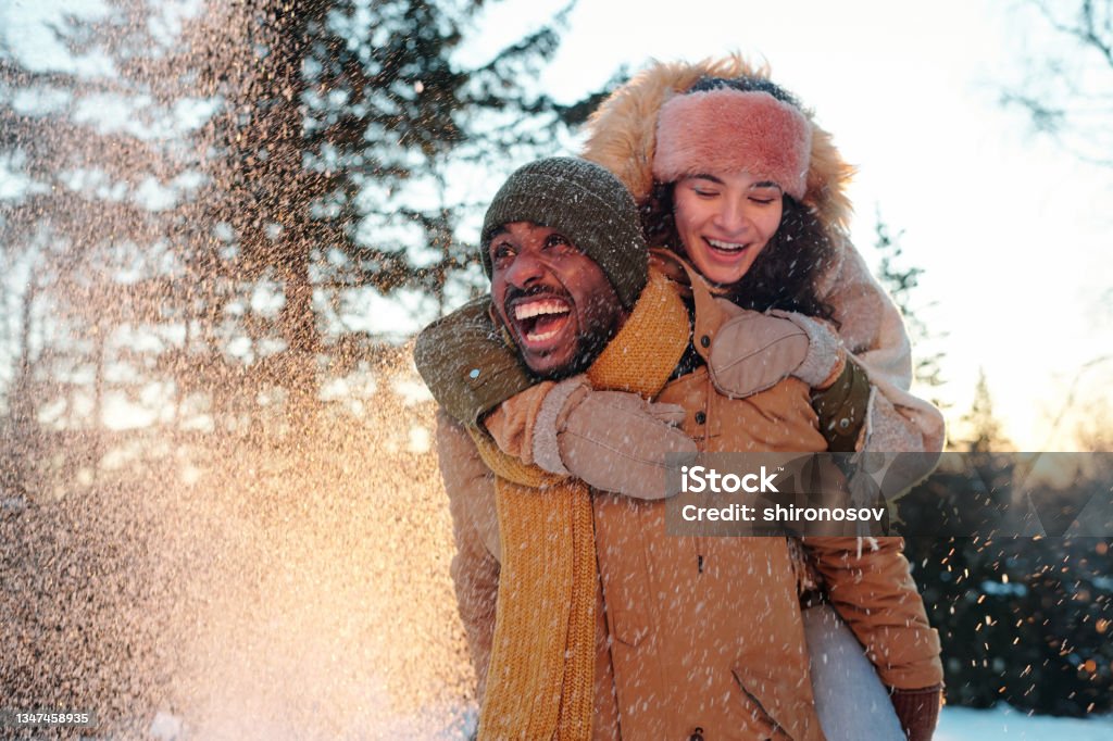 Cheerful multiracial couple in winterwear laughing while girl embracing her boyfriend Cheerful multiracial couple in winterwear laughing while girl embracing her boyfriend during snowfall Winter Stock Photo