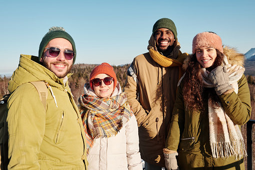 Group of young happy friends in winterwear standing in front of camera against blue sky