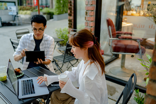 Young male and female business people having a casual work meeting in a sidewalk cafe, looking at sale charts and contracts, working on a laptop and drinking coffee