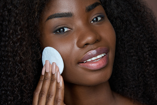 Pretty African ethnic black young woman holding cotton pad removing face make up with makeup remover cleanser on brown background. Oily skin care treatment, cleaning skincare routine concept.