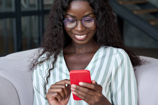 istock Young happy black African woman using mobile apps on cell phone sitting on sofa. 1347465744