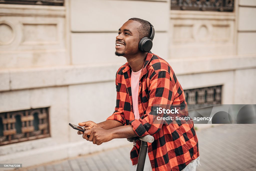 Modern man in city One man, young black male using smart phone and wearing wireless headphones while standing on scooter in city. Men Stock Photo