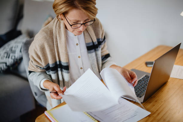 istock Mature middle aged filling in paperwork while using her laptop at home 1347467387