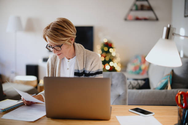 istock Mature middle aged filling in paperwork while using her laptop at home 1347468327