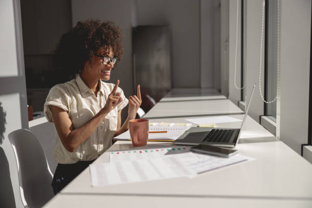 Smiling Afro Amrican female communicating in sign language online Happy pretty lady learning and communicating in sign language online while sitting in the office sign stock pictures, royalty-free photos & images