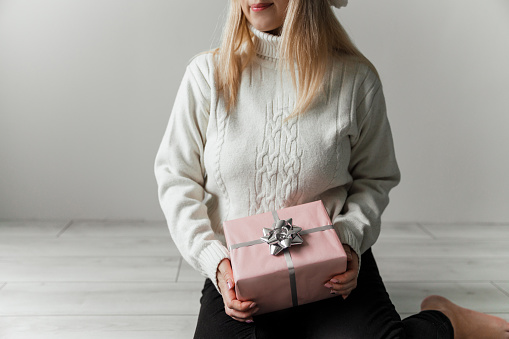 Woman in a white sweater sitting on floor and holding a pink gift in her hands on a gray background. High quality photo