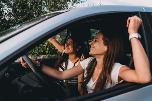 Two young adult women are enjoying a car trip together. They are sitting in the car, singing and having fun together.