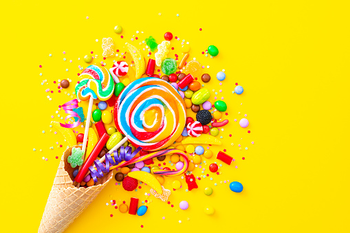 Overhead view of a waffle ice cream cone filled with multicolored candies shot on yellow background. The composition is at the left of an horizontal frame leaving useful copy space for text and/or logo at the right.
 High resolution 42Mp studio digital capture taken with SONY A7rII and Zeiss Batis 40mm F2.0 CF lens
