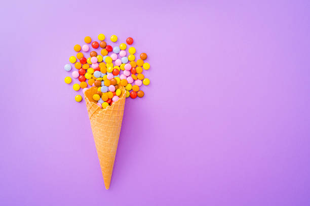 Overhead view of a waffle ice cream cone filled with multicolored candies shot on purple background. The composition is at the left of an horizontal frame leaving useful copy space for text and/or logo at the right.\n High resolution 42Mp studio digital capture taken with SONY A7rII and Zeiss Batis 40mm F2.0 CF lens