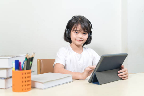 Cheerful Asian little girl student wears headphone, writes on digital tablet to study at home. Education Concept Stock Photo stock photo