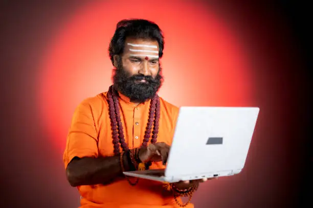 Holy Indian god man or guru with rudrakshi mala using laptop - concept of online horoscope, astrology and fortune telling using technology and internet.