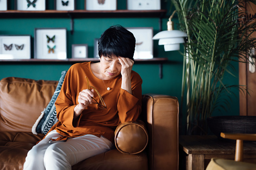 Una mujer asiática mayor angustiada que se siente mal, sufre de dolor de cabeza mientras está sentada en el sofá de la sala de estar de su casa photo
