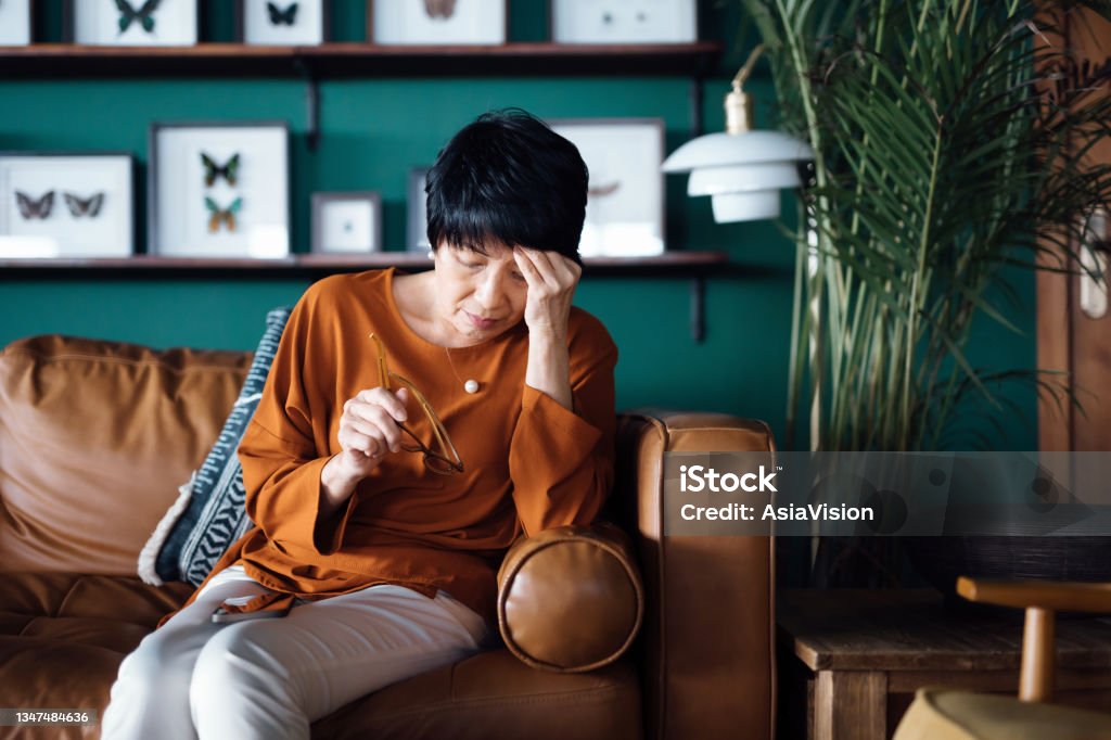 A distraught senior Asian woman feeling unwell, suffering from a headache while sitting on sofa in the living room at home Menopause Stock Photo