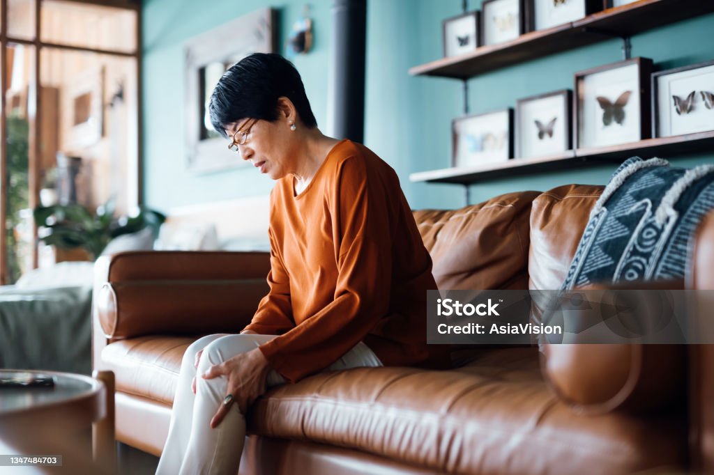 Una mujer asiática mayor angustiada que se siente mal, sufre de dolor en la pierna mientras está sentada en el sofá de la sala de estar de su casa - Foto de stock de Dolor libre de derechos
