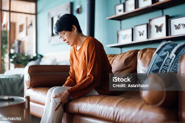 A Distraught Senior Asian Woman Feeling Unwell Suffering From Pain In Leg While Sitting On Sofa In The Living Room At Home Stock Photo - Download Image Now