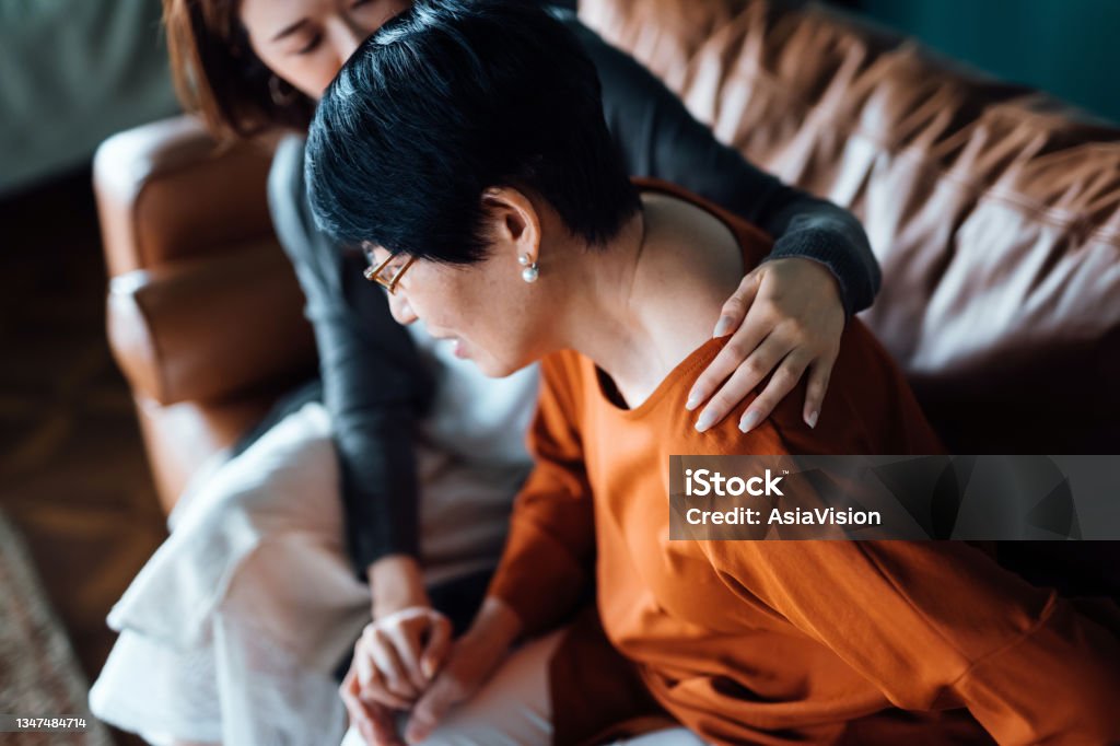 A daughter embraces her elderly mother who is feeling unwell. Holding her mother's hand giving support and consoling her while sitting on sofa in the living room at home Chronic Illness Stock Photo