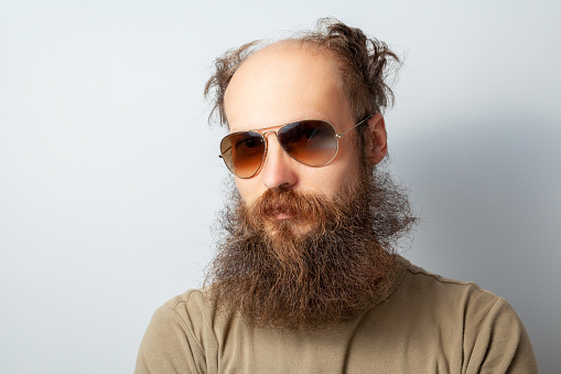 Portrait of adult handsome serious male looking at camera with calm facial expression, bald bearded man wearing T-shirt and sunglasses. Indoor studio shot isolated on gray background.