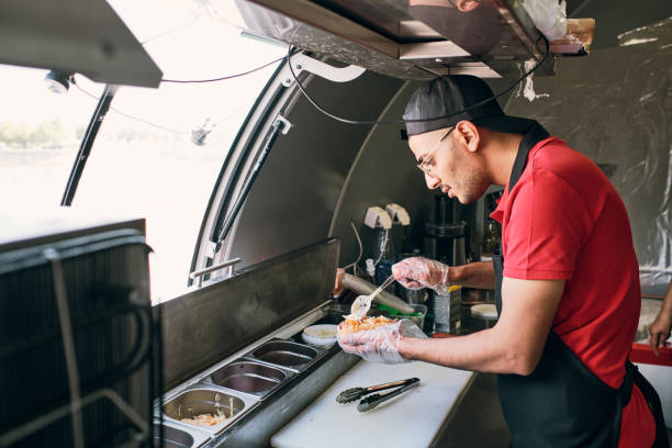 Young man in uniform cooking tasty hotdogs in food truck stock photo