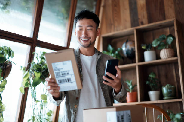 jovem asiático sorridente verificando bancos eletrônicos em seu smartphone enquanto recebia pacotes entregues de compras online em casa. compras online. banco on-line. comprando e pagando com segurança online - recebendo - fotografias e filmes do acervo