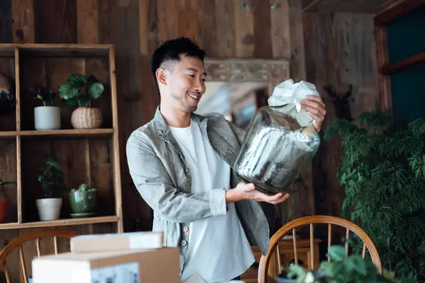 Photo of Smiling young Asian man looking happy as he unboxing the delivered package from online purchases at home, take out a glass vase. Online shopping, enjoyable customer shopping experience