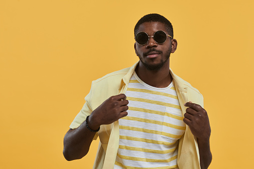 Waist up portrait of handsome African-American man wearing sunglasses and looking at camera with confidence while posing against yellow background in studio, copy space