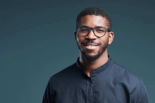 Smiling African American Man Wearing Glasses Portrait of handsome African-American man wearing glasses and looking at camera smiling while posing against deep blue background, copy space 25 year old man portrait stock pictures, royalty-free photos & images