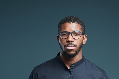 Minimal portrait of modern African-American man wearing glasses looking at camera with serious face expression while standing against deep blue background, copy space
