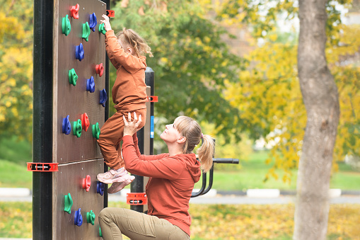 Mom helps her little daughter to overcome the artificial mountain relief on the street wall for the climbing of the sports ground. Sport and family leisure concept