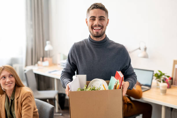 istock Young cheerful employee holding box containing office supplies 1347501199