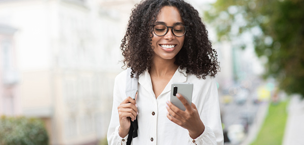 Smiling african-american student girl looking at mobile phone outdoor. Travel, people, technology, connection, mobile apps, communication and active lifestyle concept