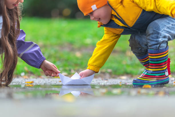 Happy kids girl and boy and colorful rubber boots playing with paper boat in puddle in autumn on nature stock photo